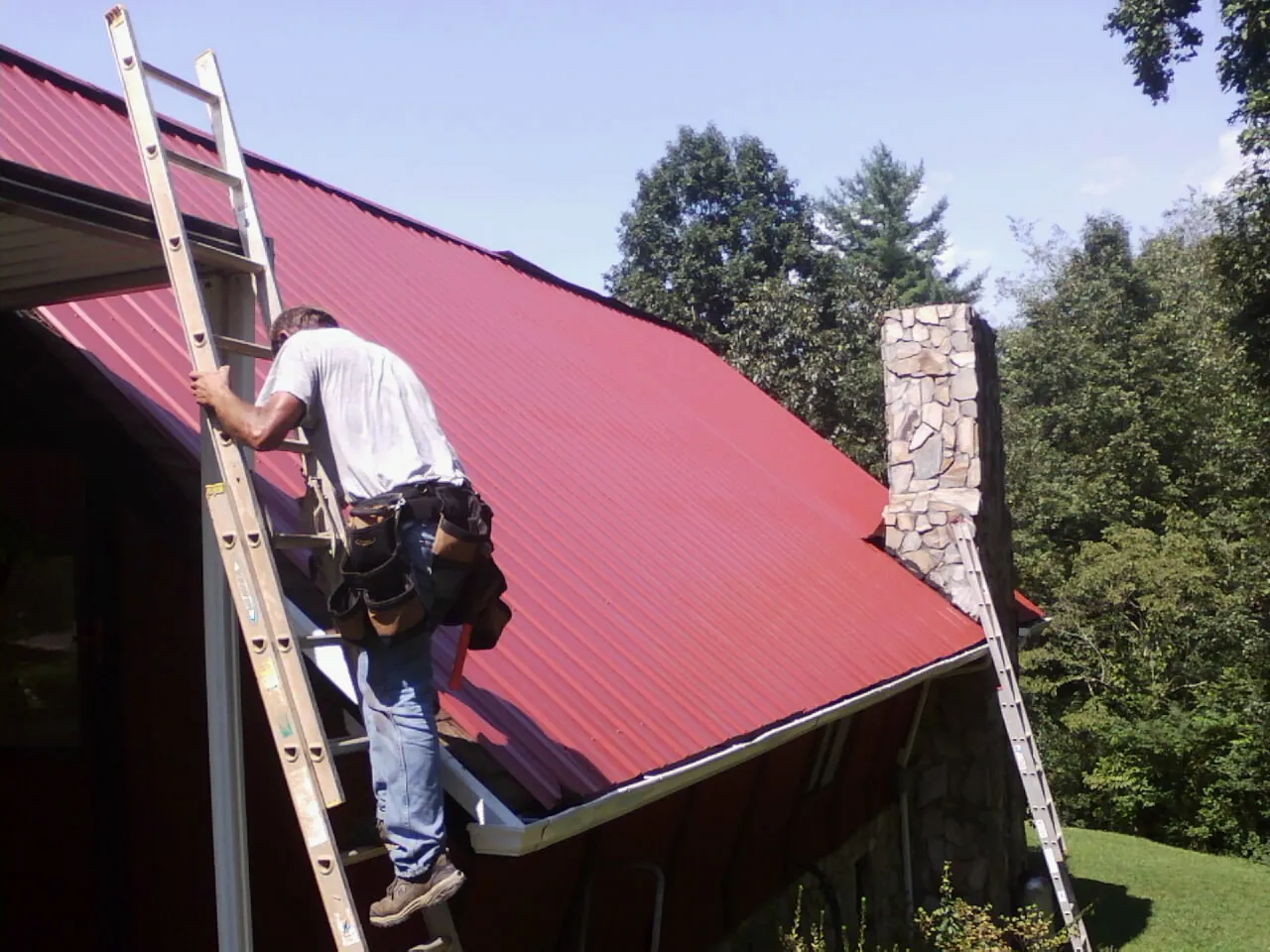 Roofer on a ladder beside a red metal roof and stone chimney — Soffit & Fascia Repair by Roofing Brothers in Bemidji
